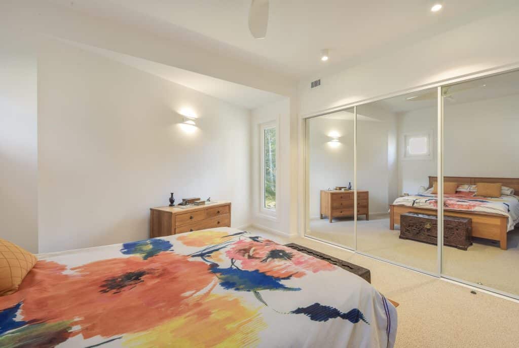 Brightly lit bedroom featuring a floral-patterned bedspread, wooden furniture, and a mirrored wardrobe, emphasizing a spacious and eco-friendly design aesthetic.