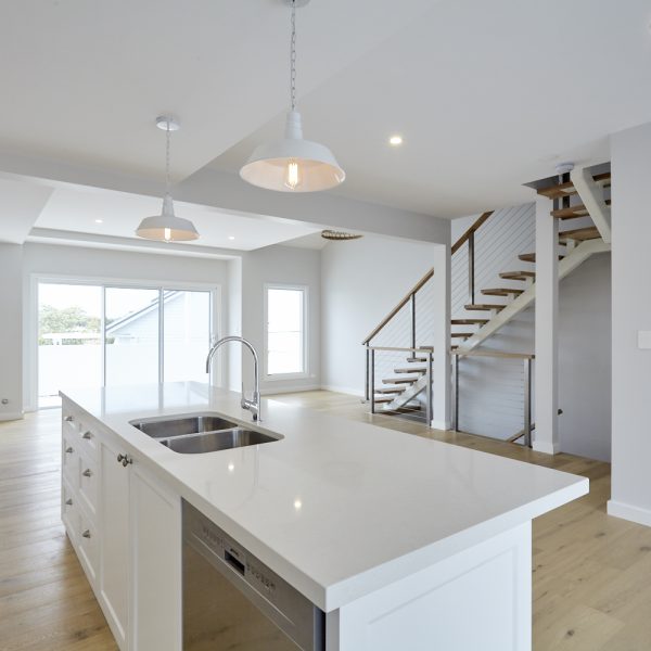 Modern kitchen interior featuring a white island with stainless steel sink, pendant lighting, and an open layout leading to a staircase, emphasizing sustainable design in the Seabreeze housing development by Living Green Designer Homes.