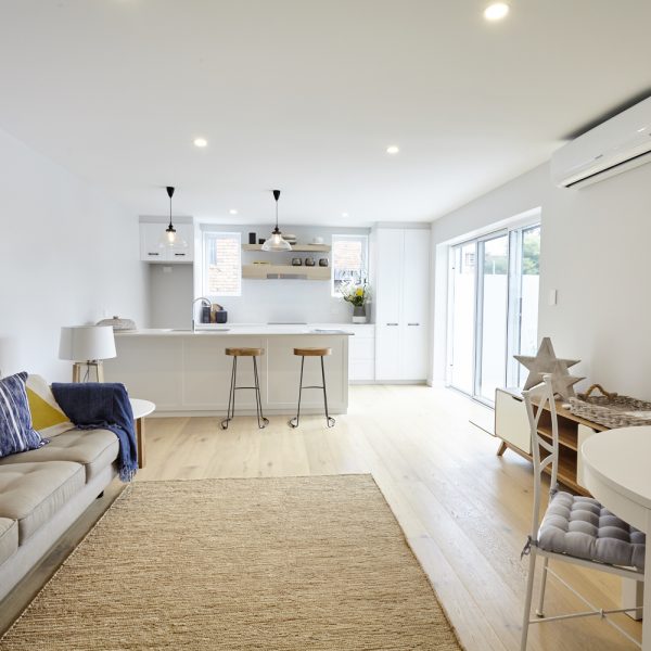 Modern open-plan living space featuring a stylish kitchen with bar stools, natural wood flooring, and bright natural light from large windows, showcasing the sustainable design of the Seabreeze project by Living Green Designer Homes.