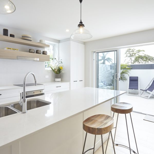 Modern kitchen with white cabinetry, quartz countertop, and wooden bar stools, showcasing a bright and open design connected to an outdoor area with lounge chairs and tropical plants, highlighting the eco-friendly features of the Seabreeze project by Living Green Designer Homes.