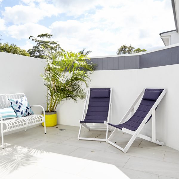 Outdoor patio area featuring white lounge chair with colorful cushions, two navy blue reclining chairs, and a potted palm tree, showcasing sustainable design elements of the Seabreeze housing development by Living Green Designer Homes.