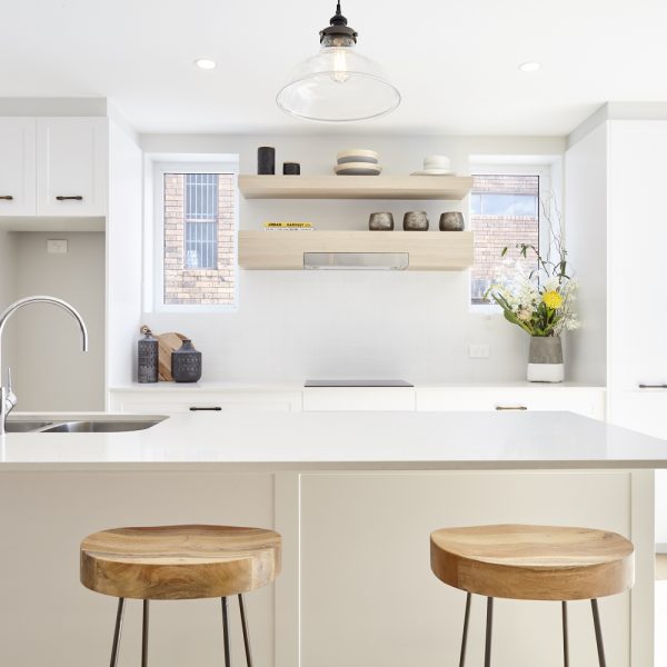 Modern kitchen design featuring a white countertop, wooden stools, minimalist cabinetry, decorative shelf with vases, and natural light from windows, highlighting the eco-friendly aesthetic of the Seabreeze project by Living Green Designer Homes.