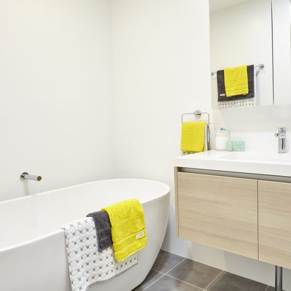 Modern bathroom featuring a freestanding bathtub, vibrant yellow and gray towels, sleek wooden cabinetry, and a contemporary mirror, showcasing the design aesthetics of the Seabreeze housing development by Living Green Designer Homes.