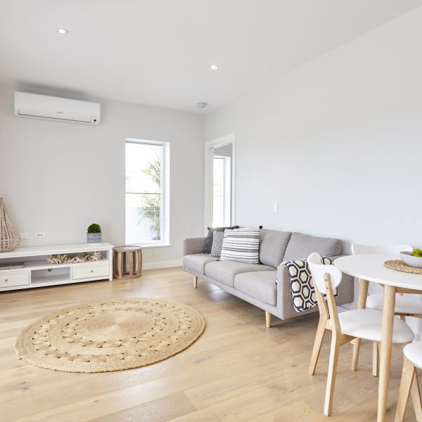 Modern living room interior featuring a gray sofa with patterned cushions, circular jute rug, wooden dining table with chairs, and large windows providing natural light, showcasing the sustainable design of the Seabreeze housing development by Living Green Designer Homes.