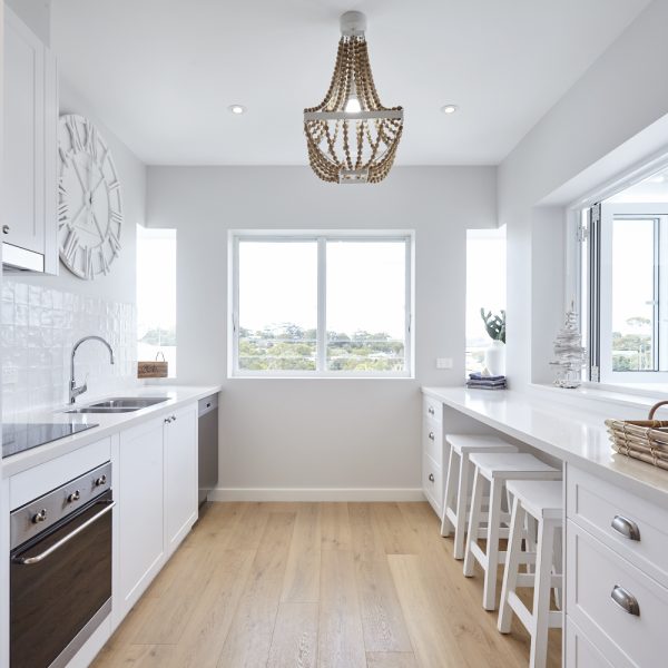 Modern kitchen design featuring white cabinetry, stainless steel appliances, and a large window with natural light, complemented by a stylish chandelier and wooden flooring, showcasing the eco-friendly aesthetics of the Seabreeze project by Living Green Designer Homes.