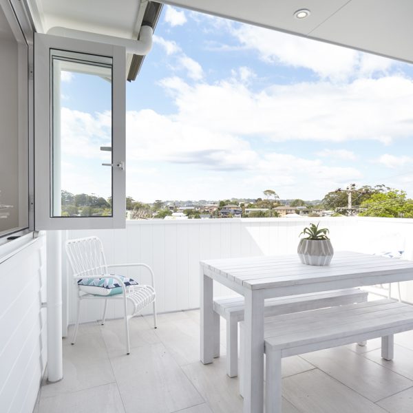 Modern outdoor patio with white wooden table and chairs, featuring a potted plant, overlooking a scenic view in the Seabreeze housing development by Living Green Designer Homes.