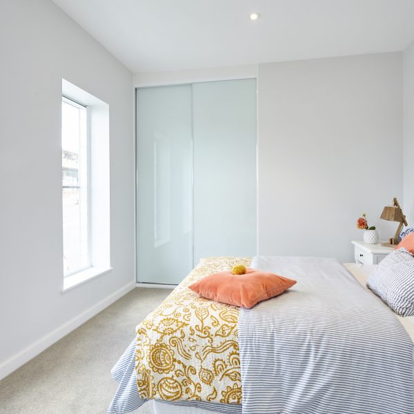 Bright bedroom in Seabreeze housing development featuring a bed with patterned throw, orange pillow, large window, and modern sliding wardrobe, showcasing eco-friendly design by Living Green Designer Homes.