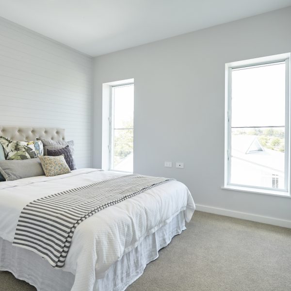 Modern bedroom design featuring a plush bed with decorative pillows, striped blanket, and large windows providing natural light, showcasing the sustainable and stylish interiors of the Seabreeze housing development by Living Green Designer Homes.