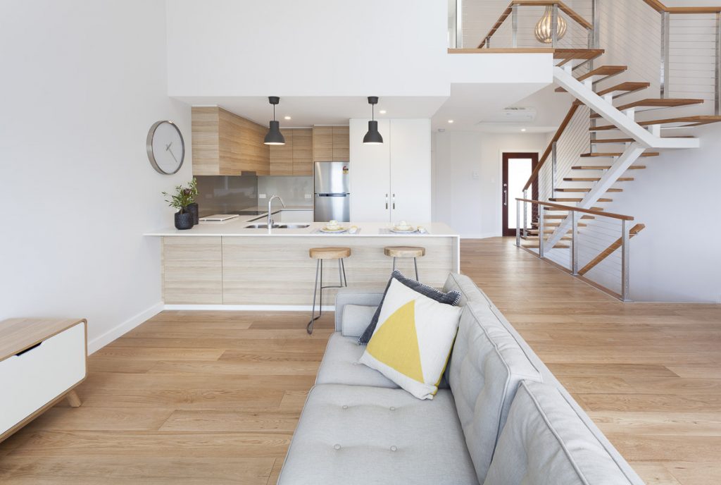 Modern open-plan living space featuring a stylish kitchen with wooden cabinetry, sleek appliances, and a cozy sofa adorned with a yellow and gray pillow, emphasizing sustainable design and contemporary architecture by Living Green Designer Homes.