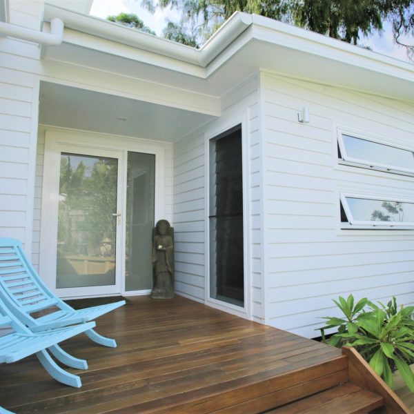 Family-friendly home entrance featuring light blue chairs, wooden decking, modern architecture, and a decorative statue, reflecting sustainable design principles by Living Green Designer Homes.