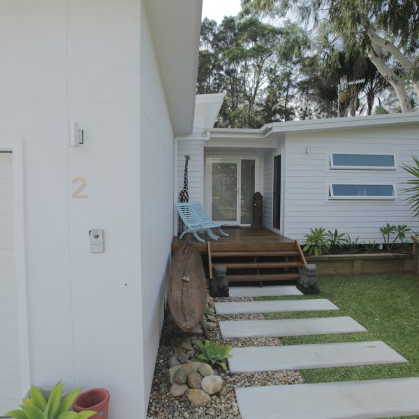 Modern family-friendly home entrance with wooden deck, blue chairs, stone pathway, and landscaped garden, showcasing sustainable design features by Living Green Designer Homes.