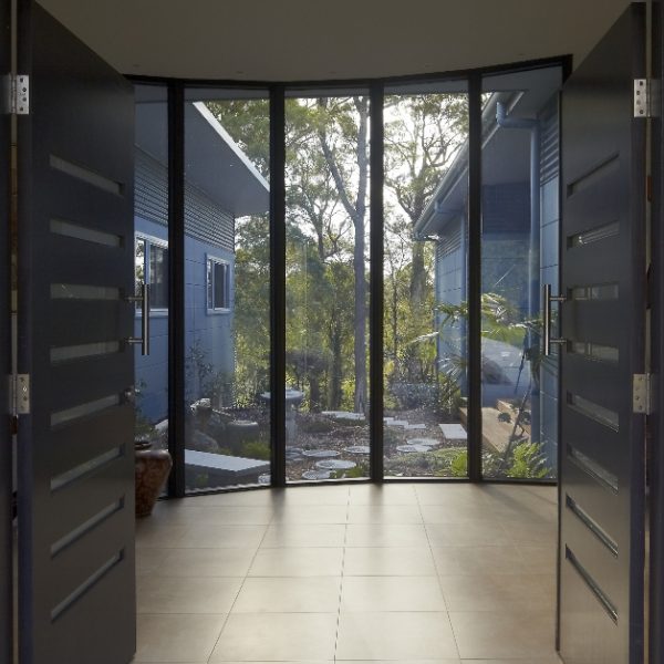 Modern entrance with double doors opening to a serene outdoor space featuring lush greenery and a stone pathway, highlighting the sustainable design of the Hilltop Hideaway home by Living Green Designer Homes.