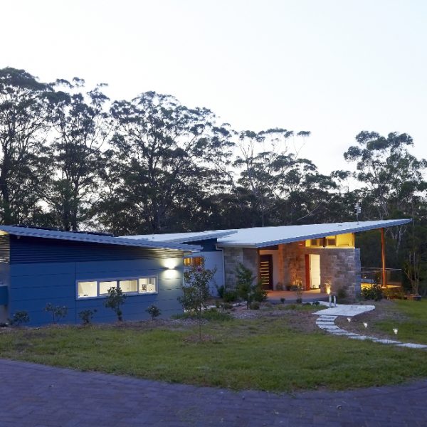 Modern sustainable home design featuring a blue exterior, stone accents, and a unique roof structure, surrounded by greenery, showcasing the 'Hilltop Hideaway' project by Living Green Designer Homes.