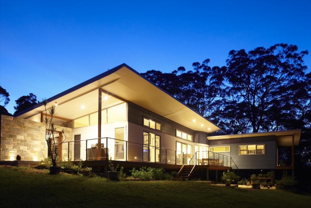 Modern sustainable home design with large windows, illuminated at dusk, surrounded by greenery, showcasing energy-efficient construction by Living Green Designer Homes.