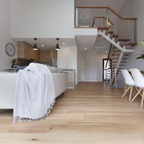 Modern open-plan living space in Seabreeze housing development featuring wooden flooring, a cozy sofa with a gray throw, stylish staircase, and contemporary kitchen design by Living Green Designer Homes.