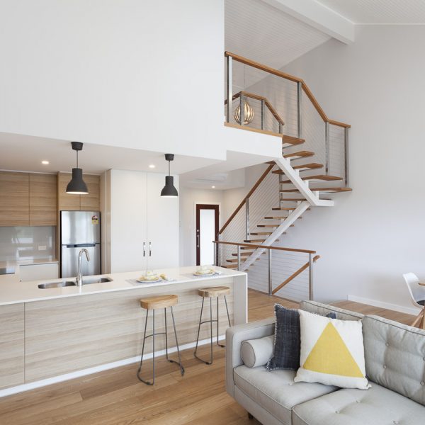 Modern kitchen and living area in Seabreeze housing development by Living Green Designer Homes, featuring wooden cabinetry, sleek appliances, and a staircase with stylish railing.
