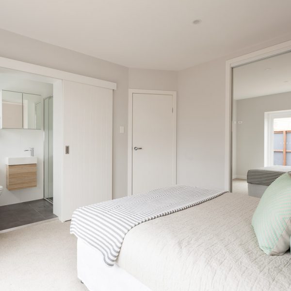 Modern bedroom interior featuring a bed with striped throw, adjacent bathroom with sliding door, and natural light illuminating the space, showcasing the design and quality of the Seabreeze housing development by Living Green Designer Homes.