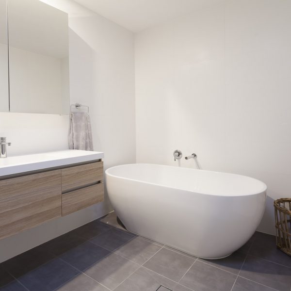 Modern bathroom design featuring a freestanding white bathtub, minimalist wooden cabinetry, and a large mirror, emphasizing sustainable and stylish interior elements from the Seabreeze project by Living Green Designer Homes.