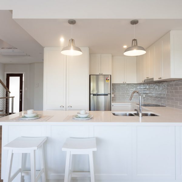 Modern kitchen interior featuring a white island with two stools, stylish pendant lighting, stainless steel appliances, and a sleek backsplash, showcasing the design elements of the 'Seabreeze' housing development by Living Green Designer Homes.