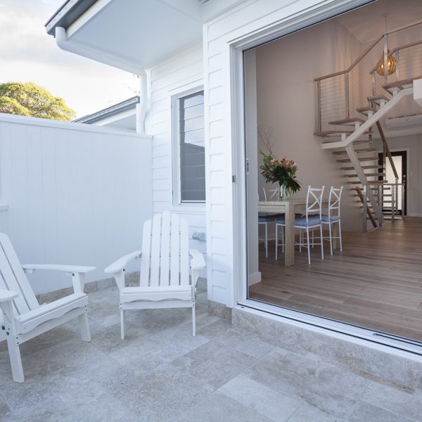 Outdoor seating area with white Adirondack chairs and a view into a modern interior featuring a staircase and dining table, highlighting the design of the Seabreeze project by Living Green Designer Homes.