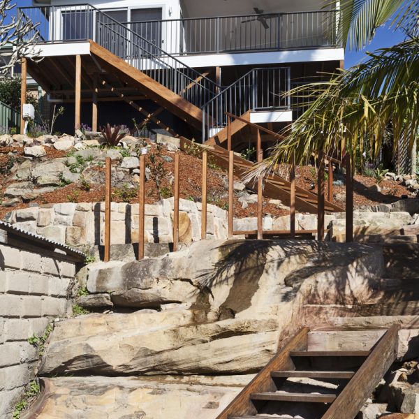 Sustainable home design featuring wooden stairs leading to a modern elevated house with a landscaped rock garden, showcasing Living Green Designer Homes' innovative construction techniques.
