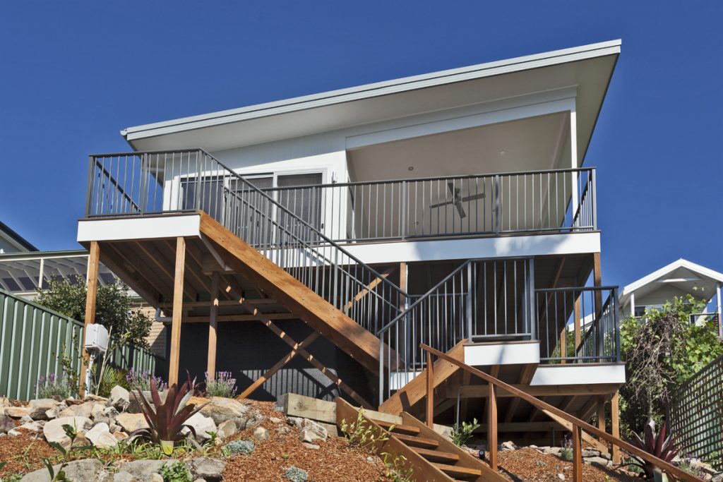 Modern sustainable home with wooden deck and railing, set on a landscaped hillside, showcasing energy-efficient design by Living Green Designer Homes.