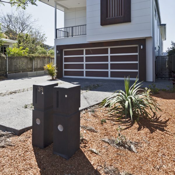 Modern sustainable home exterior with stylish design, featuring a two-tone garage door, house number 39, and contemporary letterboxes labeled A and B, surrounded by landscaped garden with mulch and greenery.